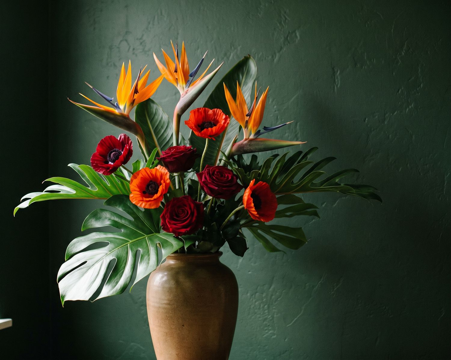 Strauß aus orangen Strelitzien, rotem Mohn, dunkelroten Rosen und Monstera-Blättern in einer Tonvase vor einer dunkelgrünen Wand